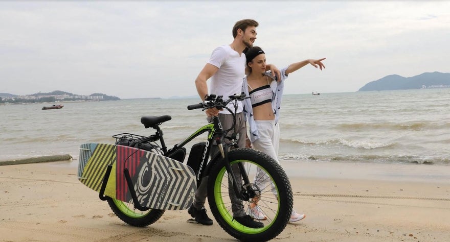 A man and woman walking along the beach. The man has his arm around the woman and is steering his bike with the other. The woman is leaning into him and pointing into the distance.