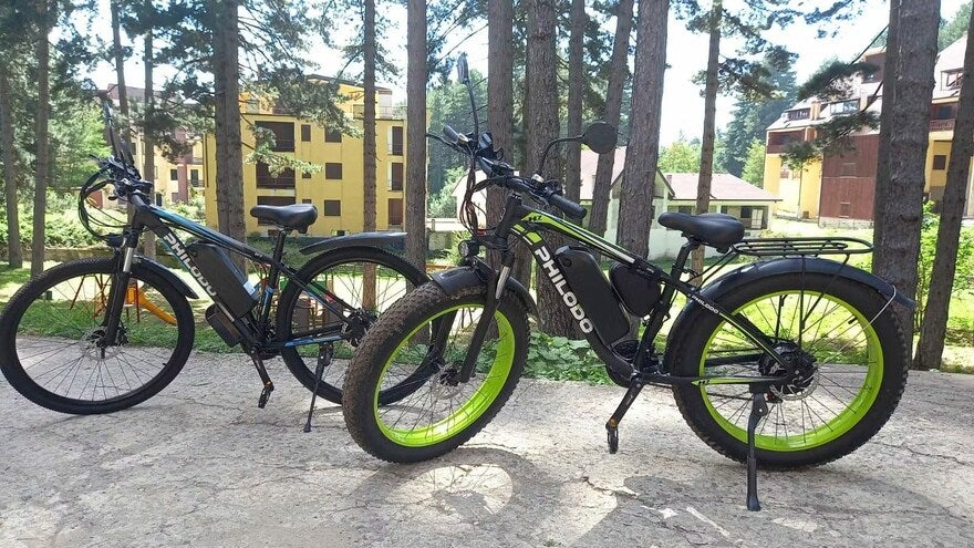 Two bikes on a path in front of trees with some buildings in the background.